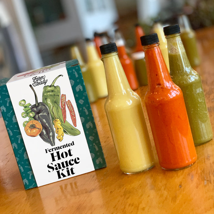 On a wooden table rests a Fermented Hot Sauce Kit from FarmSteady, with three bottles of yellow, orange, and green homemade hot sauces. More bottles in the background hint at an umami depth thats truly tantalizing.