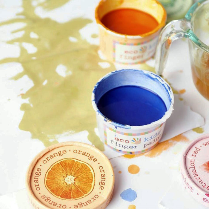Open containers of eco-kids Finger Paint Set in blue and orange rest on a white surface with paint splatters. Nearby are lids with orange text. A glass measuring cup is partially visible, highlighting these all-natural, eco-friendly paints as fun and food-safe for creative minds.