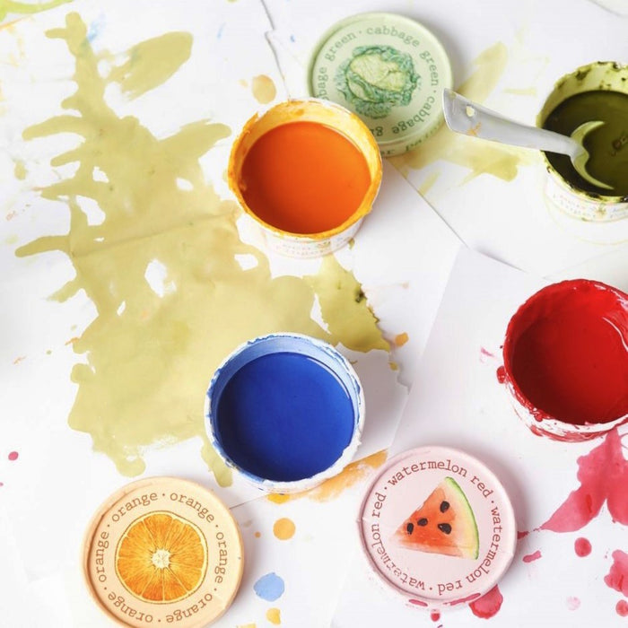 Open jars of eco-kids Finger Paint Set in yellow-green, orange, blue, and red are on a splattered white surface. Nearby are lids with illustrations of an orange, watermelon, and cabbage. A small metal spoon is in the green paint.