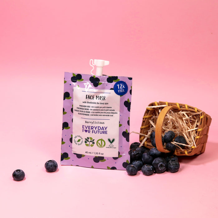 A Firming Blueberry Face Mask by Everyday For Future, with a Berrylicious design featuring blueberry illustrations, sits on a pink surface. Next to it is a small wicker basket with straw and scattered blueberries. The vegan mask offers antioxidants and can be used 12 times.