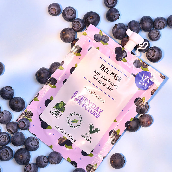A purple pouch of Everyday For Futures Firming Blueberry Face Mask, with antioxidant properties, is surrounded by fresh blueberries on a light blue background. The packaging features blueberry illustrations and a 100% vegan label.