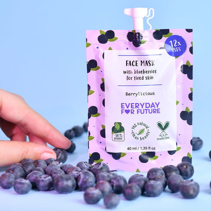 A hand holds a sachet of Everyday For Futures Firming Blueberry Face Mask, encircled by fresh blueberries. The pink pouch, adorned with a blueberry pattern, revitalizes tired skin. It features 50% plastic-free packaging and is vegan and cruelty-free, offering 12 antioxidant-rich uses.