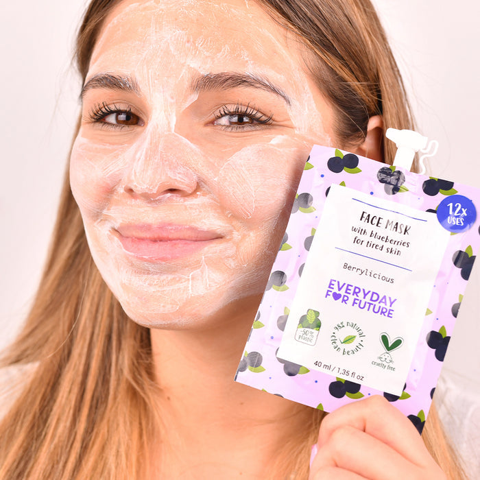 A person with long hair applies a vegan face mask from a purple and white Berrylicious pouch by Everyday For Future. This Firming Blueberry Face Mask, rich in antioxidants, rejuvenates tired skin and provides 12 soothing uses.