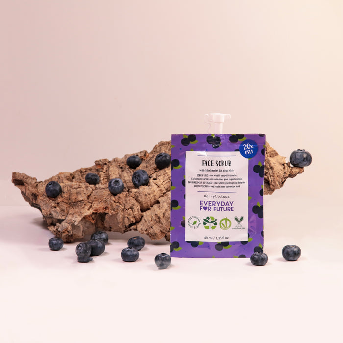 A Firming Blueberry Face Scrub from Everyday For Future stands upright on a light surface, surrounded by scattered blueberries and bark. It boasts antioxidant properties, with purple packaging featuring green illustrations and white text/icons against a plain background.