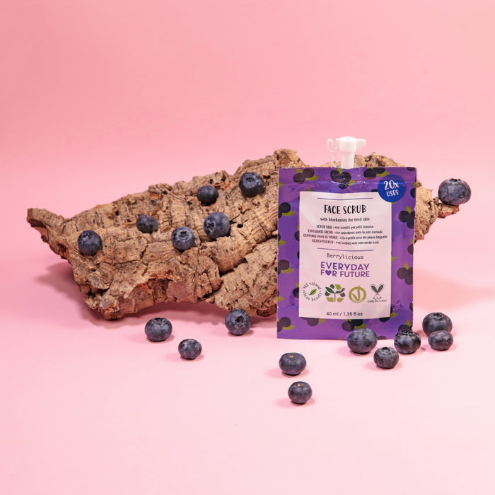 Introducing Everyday For Futures Firming Blueberry Face Scrub: a pouch with a purple label showcasing vibrant blueberries on a pink backdrop, nestled among fresh blueberries and tree bark, featuring vegan-friendly, antioxidant properties for naturally radiant skin.