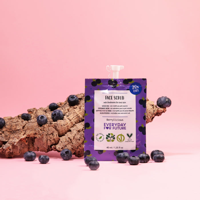 A pouch of Firming Blueberry Face Scrub by Everyday For Future sits amidst scattered blueberries and tree bark on a pink backdrop. The package emphasizes its antioxidant and vegan features, appealing to eco-conscious skincare fans seeking a burst of berry goodness.
