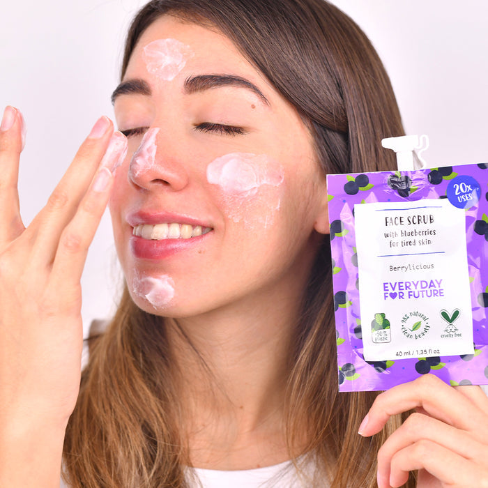 A woman uses the creamy Firming Blueberry Face Scrub from the purple and white Berrylicious package. Perfect for tired skin with antioxidants, it features eco-friendly symbols, offers up to 20 uses, and highlights a vegan formula, all under the brand Everyday For Future.