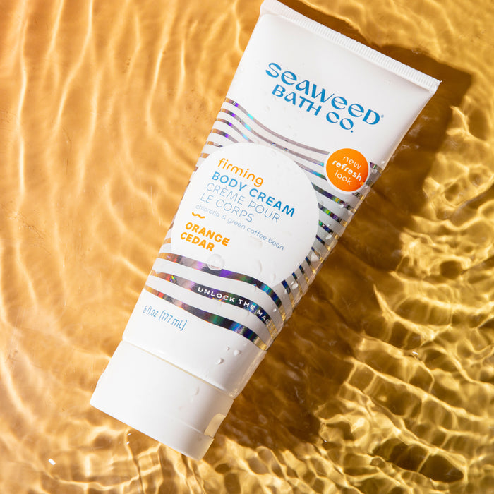A tube of The Seaweed Bath Cos Firming Body Cream in orange cedar scent glides on shimmering golden water, enriched with bladderwrack seaweed for rejuvenation. It features a colorful wave design and new packaging label.