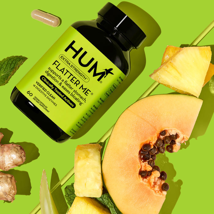 A black bottle of HUM Nutritions Flatter Me Extra Strength - For Bloat Relief rests on a yellow background, surrounded by papaya, pineapple, ginger, parsley slices, and a capsule. The digestive enzyme blend supports debloating for a flatter tummy. The green label features yellow and black text.