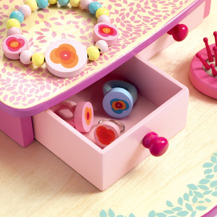 A small pink drawer opens to reveal colorful rings with floral designs, while a matching bracelet rests atop. This charming scene is set on DJECOs Flora Dressing Table Play Set, crafted from 100% FSC® Certified non-toxic wood adorned with floral patterns.