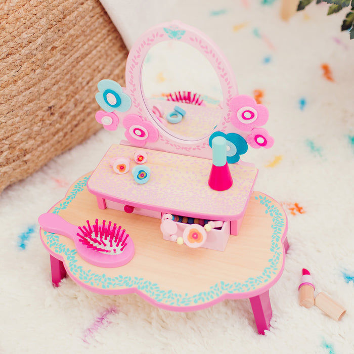 A charming Flora Dressing Table Play Set by DJECO sits on a soft rug, featuring a pink table with mirror, flower decorations, and mini cosmetics. Made from 100% FSC® Certified Wood, its playful pink, blue, and white accents inspire imaginative play with non-toxic materials.
