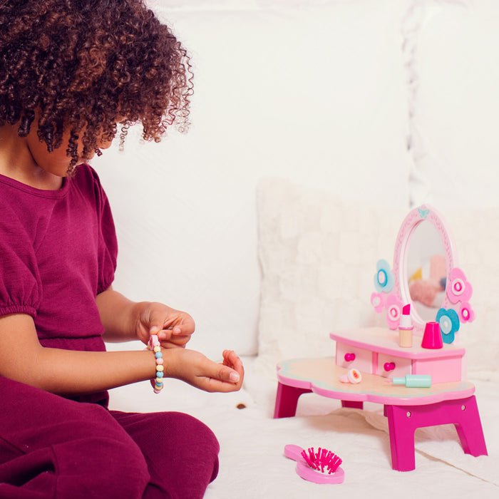 A child with curly hair in a maroon shirt sits on a white bed, examining a colorful beaded bracelet. In front of them is the DJECO Flora Dressing Table Play Set, a pink toy vanity with mirror and creative, non-toxic accessories for imaginative play, made from 100% FSC® Certified Wood.