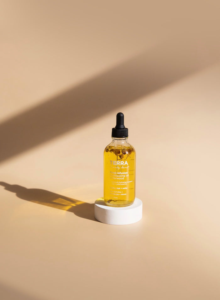 The Floral Infusion Multitasking Oil for Body + Hair by Terra Beauty Bars sits on a small white circular stand against a beige background. The dropper bottle contains a golden liquid with the serene allure of Jasmine Scent, casting a soft shadow to the left, embodying tranquility.