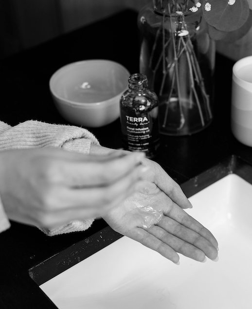 In a black and white image, someone in a knit sweater pours Floral Infusion Multitasking Oil for Body + Hair from Terra Beauty Bars into their palm. Nearby, a bowl, flowers in a vase with jasmine scent, and a cup are on the counter beside a square sink.