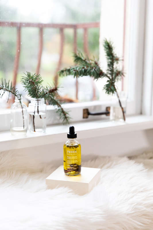 A bottle of Terra Beauty Bars Floral Infusion Multitasking Oil for Body + Hair, with natural oils, rests on a white cube atop a fluffy white surface by the window. Small vases of pine branches adorn the sill, creating a serene and natural ambiance to enhance your self-care routine.