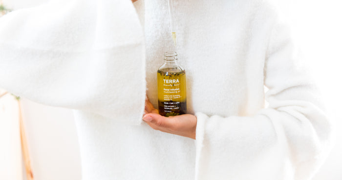 A person in a white fluffy sweater holds a clear bottle labeled Terra Beauty Bars, filled with yellow multitasking oil called Floral Infusion, ideal for body and hair. Enhanced with natural oils, the dropper-capped bottle adds serenity to any self-care routine against a softly blurred background.