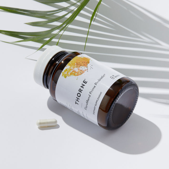 A dark brown FloraMend Prime Probiotic® bottle by Thorne rests on a white surface with two capsules nearby. Shadows from a green palm leaf add a natural touch, suggesting benefits for gut health and weight management.