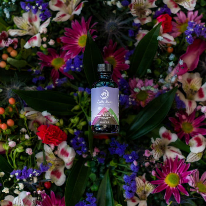 A bottle labeled Flow Your Own Way by Little Moon Essentials, infused with essential oils, stands amidst a vibrant display of colorful flowers including red, purple, and pink blossoms, framed by lush green leaves for perfect monthly relief.
