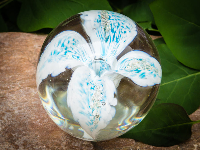 Clear glass flower paperweight with blue and white swirled petals encasing cremation ashes on a stone surface.