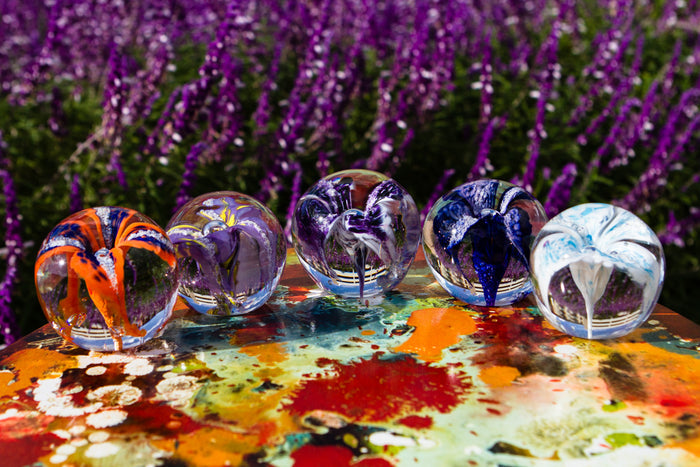 Row of colorful glass flower paperweights arranged on a paint splattered table outdoors.