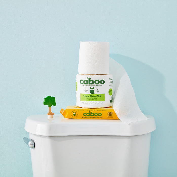 A package of Caboos Flushable Wipes rests on a toilet tank lid next to its yellow packaging box. The wipes are partially pulled out, and a small green tree-shaped ornament is nearby. The wall is painted pastel blue.