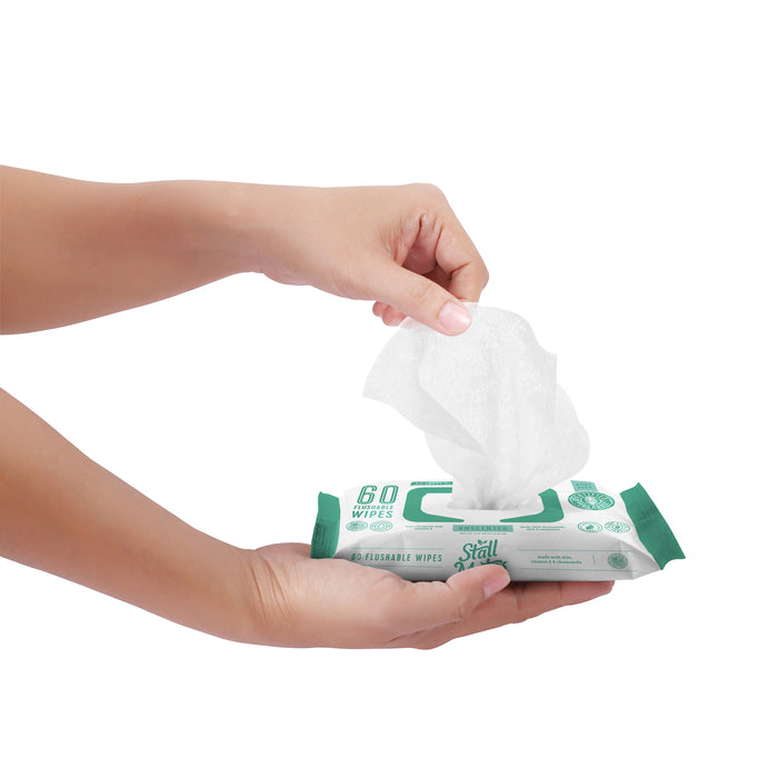 Someone grabs a Stall Mates Flushable Wipe from a green and white pack marked 60 wipes, featuring Hydraspun Dispersible tech. The design and text emphasize hypoallergenic benefits against a crisp white backdrop, showcasing its convenience and care.