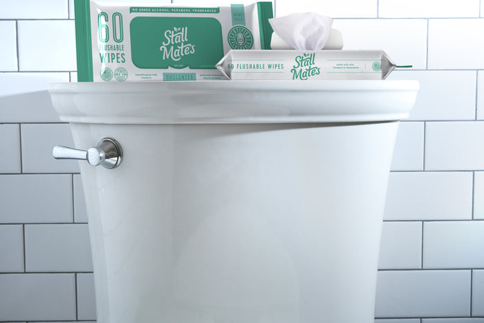 A white toilet tank against a white tile wall holds two packs of Stall Mates Flushable Wipes. One pack, labeled 60 Flushable Wipes, stands upright, while the other is open to reveal a wipe made with Hydraspun Dispersible technology.
