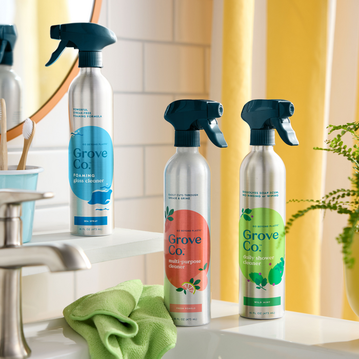 Three Grove Co. recyclable aluminum spray bottles labeled Multi-Purpose Cleaner (VIP Discount), foaming glass cleaner, and daily shower cleaner sit on a bathroom counter with mirrors and tiles, accompanied by a green cloth and toothbrush holder, ready to tackle any household mess.