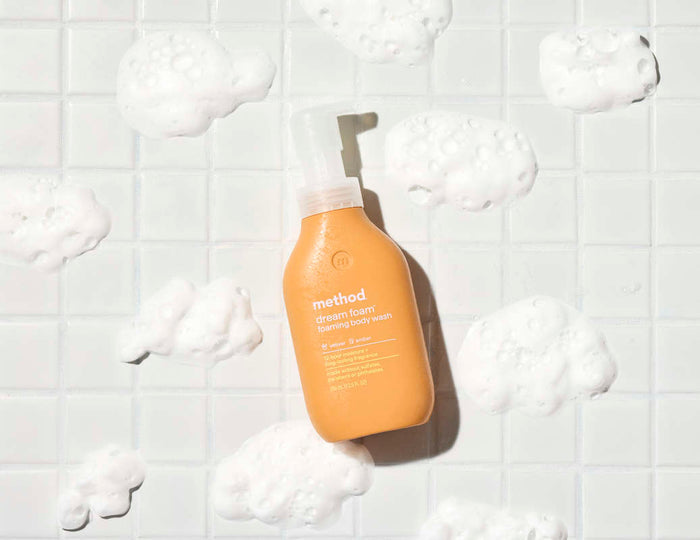 A Method Foaming Body Wash bottle, made from recycled plastic, rests on white tiles surrounded by foam clusters. This cruelty-free formula features an orange bottle with a white pump.