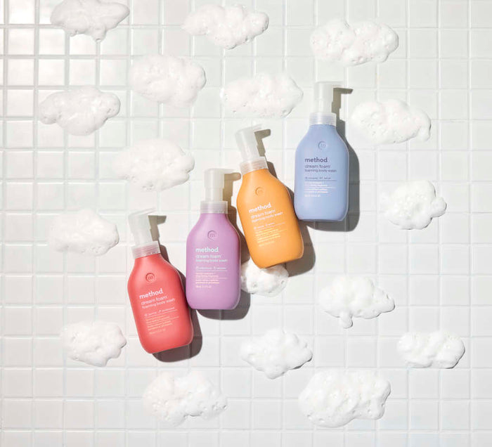 Four Method Foaming Body Wash bottles—pink, purple, orange, and blue—against a white tiled backdrop with foamy bubbles like clouds. Made from recycled plastic, these eco-friendly bottles add color and sustainability to your space.