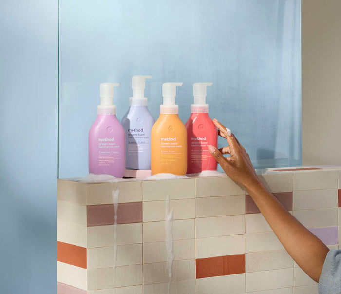 On a tiled ledge, four vibrant method Foaming Body Wash bottles from recycled plastic are lined up, each with a pump. A hand reaches for one amidst frosted glass and beige, pink, and brown tiles. Soap suds cover the surface.