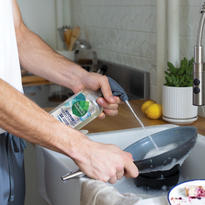 In the kitchen, someone uses Seventh Generations Foaming Dish Spray to wash a frying pan under running water. The countertop displays a plant, sponge, and lemons.
