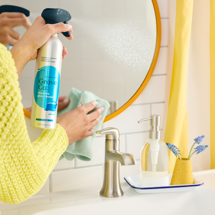 In a bathroom with a faucet, soap dispenser, and lavender in a vase on the counter, someone in a yellow sweater uses Grove Co.s Foaming Glass Cleaner from a recyclable aluminum bottle to spray onto a mirror. They wipe it with a green cloth, making the orange-framed mirror shine streak-free.