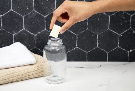 A hand places a Meliora Foaming Hand Soap Refill Tablet into a clear glass jar on a marble surface. Two folded towels, one white and one beige, are stacked beside it against hexagonal tiles, embracing the elegance of plastic-free packaging.