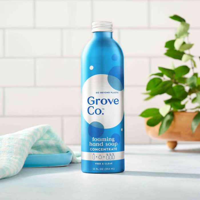 A blue bottle of Grove Co. Foaming Hand Soap Refill with plant-based ingredients sits on a countertop. Its label reads Go beyond plastic and Free & Clear. In the background, a plant thrives alongside a cloth and soap dish, highlighting sustainable packaging.