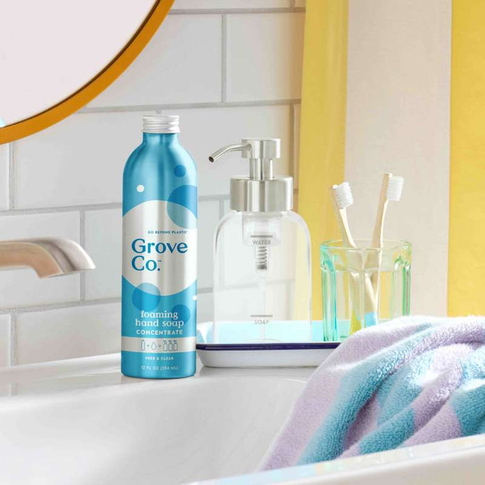 On a bathroom counter sits a blue bottle of Grove Co. Foaming Hand Soap Refill with plant-based ingredients and sustainable packaging. Nearby, a glass soap dispenser, two toothbrushes in a green holder, and a fluffy towel are set beneath the mirror and yellow curtain.