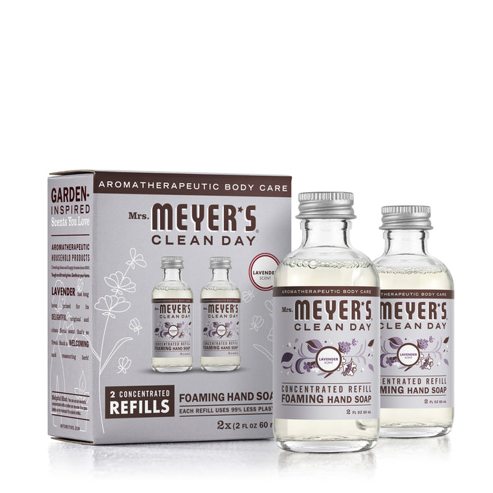 Two glass bottles of Mrs. Meyers Foaming Hand Wash Concentrate Refills in Lavender, made with natural cleaning agents, sit beside their packaging box adorned with lavender images and product details that highlight garden-inspired scents and aromatherapeutic benefits.