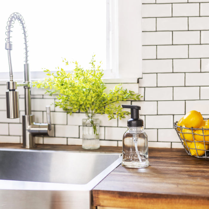 A kitchen sink with a stainless steel faucet stands before a white subway tile backsplash. Nearby, theres a small plant in a jar, Mrs. Meyers Foaming Hand Wash Concentrate Refills, and a wire basket of lemons on the wooden countertop.