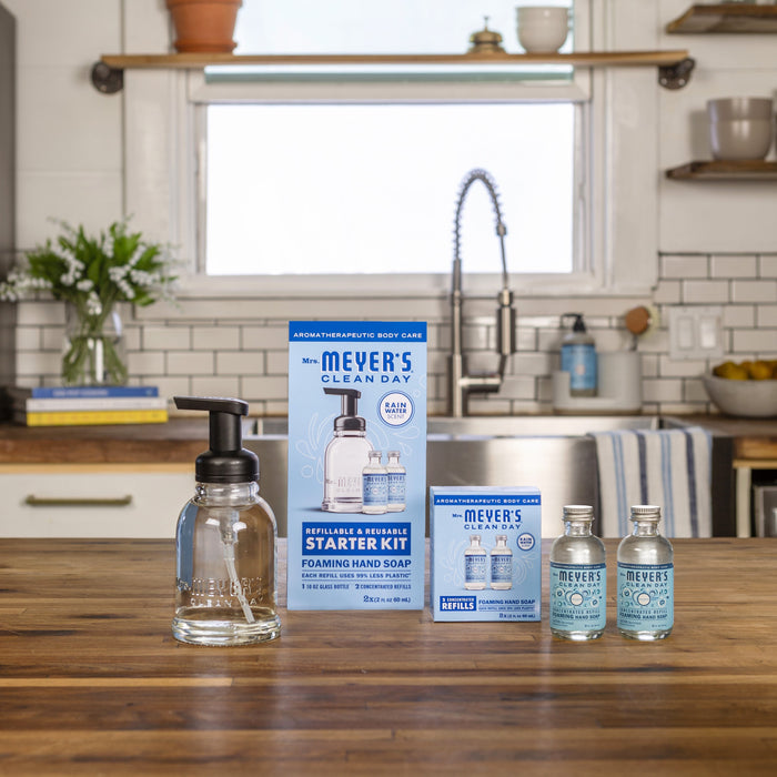 A kitchen scene showcases Mrs. Meyers Foaming Hand Wash Concentrate Starter Set on a wooden countertop, featuring a starter kit box and two refill bottles, along with a glass dispenser with a black pump. A sunlit window and potted plant add charm to this natural cleaning agent setting.