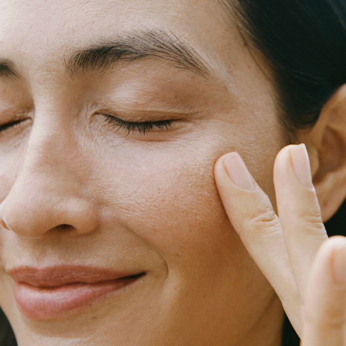 A close-up of a person with smooth skin and a soft smile, eyes closed, gently touching their cheek—highlighting the visible results of Ursa Majors Forest Alchemy Eye Cream for reducing fine lines.