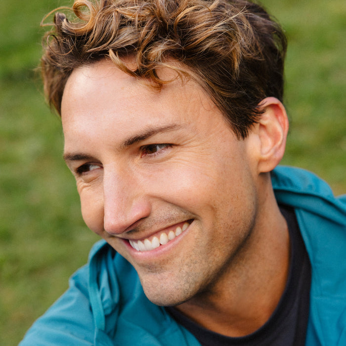 A young man with wavy light brown hair smiles and looks to the side, appearing refreshed with no dark circles or fine lines—thanks to Ursa Major’s Forest Alchemy Eye Cream—against a blurred green outdoor background.