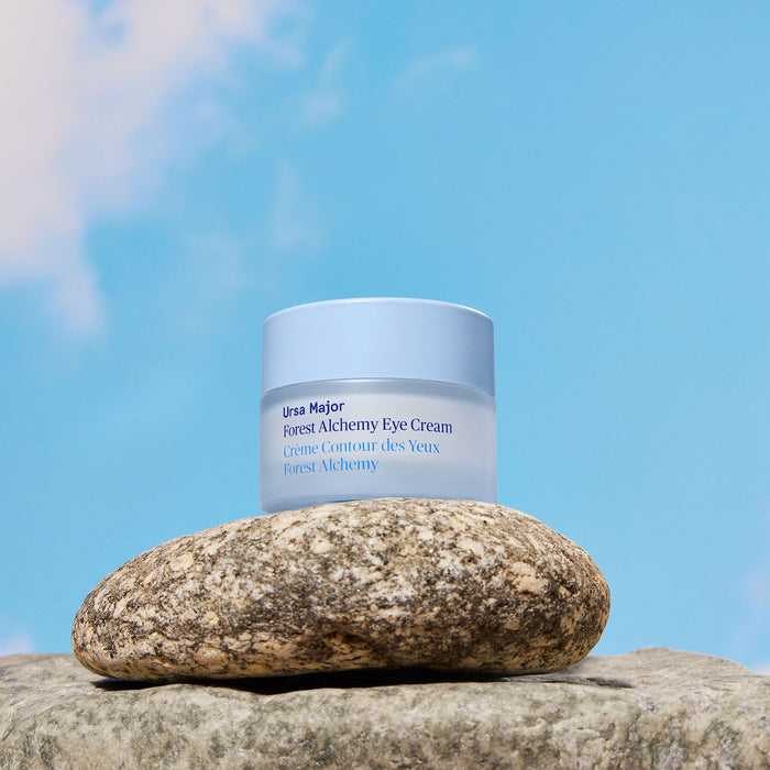 A jar of Ursa Major Forest Alchemy Eye Cream, designed to address dark circles and fine lines, rests on a large stone beneath a blue sky scattered with white clouds.