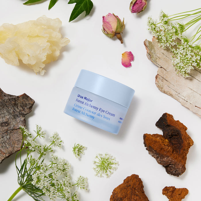 A light blue jar of Ursa Major Forest Alchemy Eye Cream, designed to target fine lines and dark circles, sits on a white surface surrounded by dried flowers, tree bark, and white blossoms in a botanical arrangement.