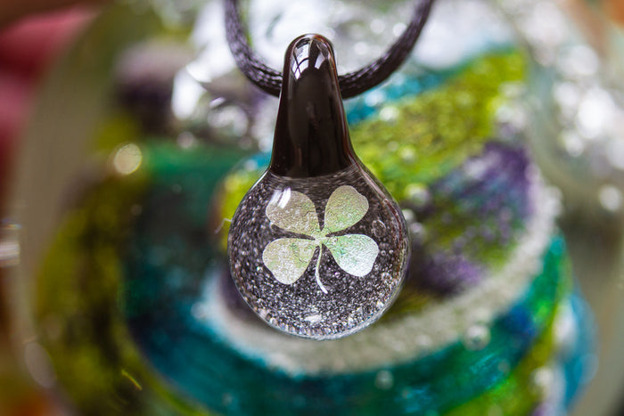 Close-up of a glass teardrop pendant with a holographic four leaf clover and cremation ash on a black adjustable cord.