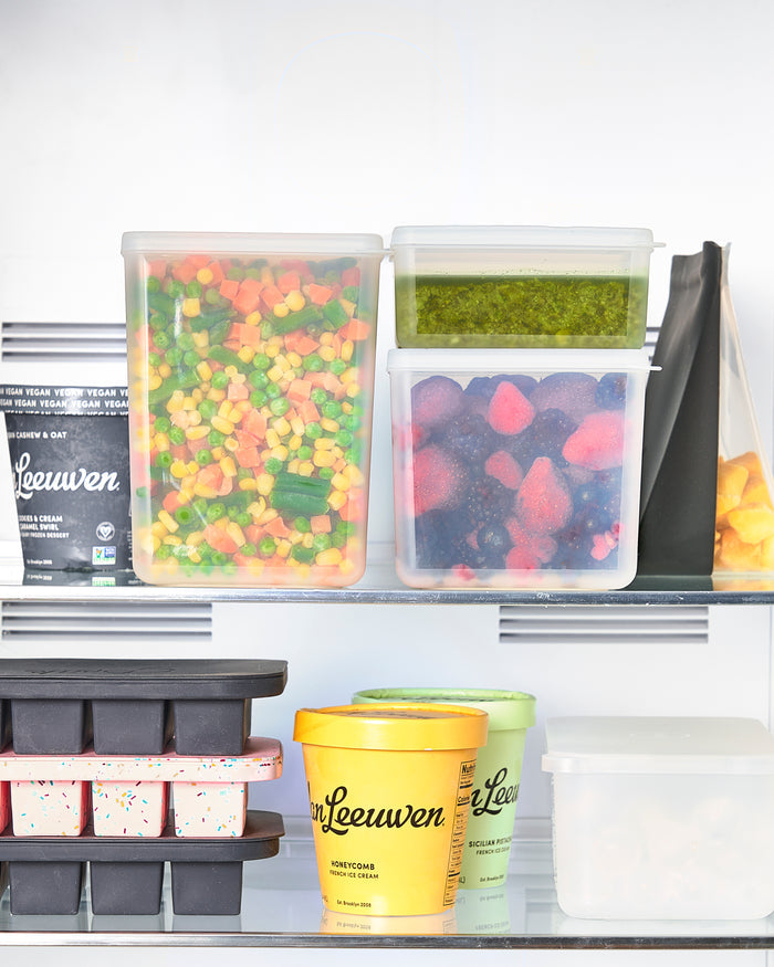 The fridge contains assorted frozen foods like packed vegetables, berries, ice cream tubs, and W&Ps black stackable Freezer Cubes. The background features other neatly arranged frozen items for sustainable storage.