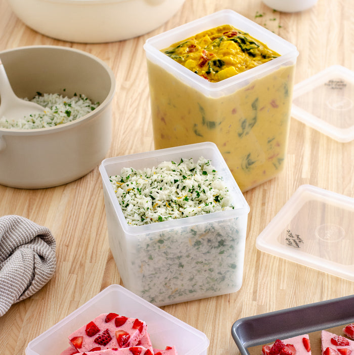 Three Freezer Cubes by W&P containing yellow curry with vegetables, cilantro rice, and a strawberry dessert sit on a wooden surface. Nearby is a beige bowl of rice and a gray cloth, highlighting this sustainable storage for your culinary creations.