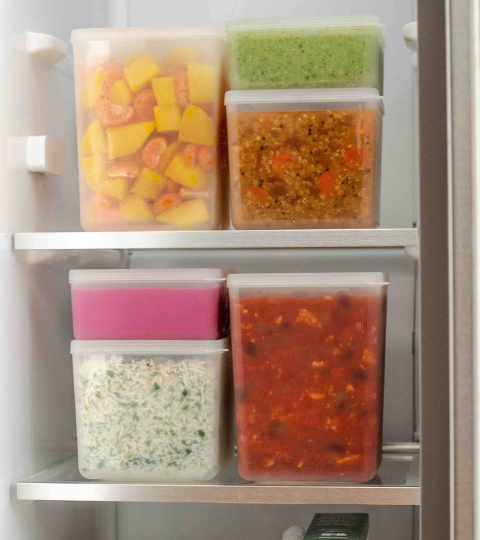 W&Ps Freezer Cubes, stackable and clear, brim with vibrant foods like fruit and vegetable mix, green sauce, mixed grains, and herb rice. These sustainable storage solutions maximize efficiency and freshness on two refrigerator shelves.