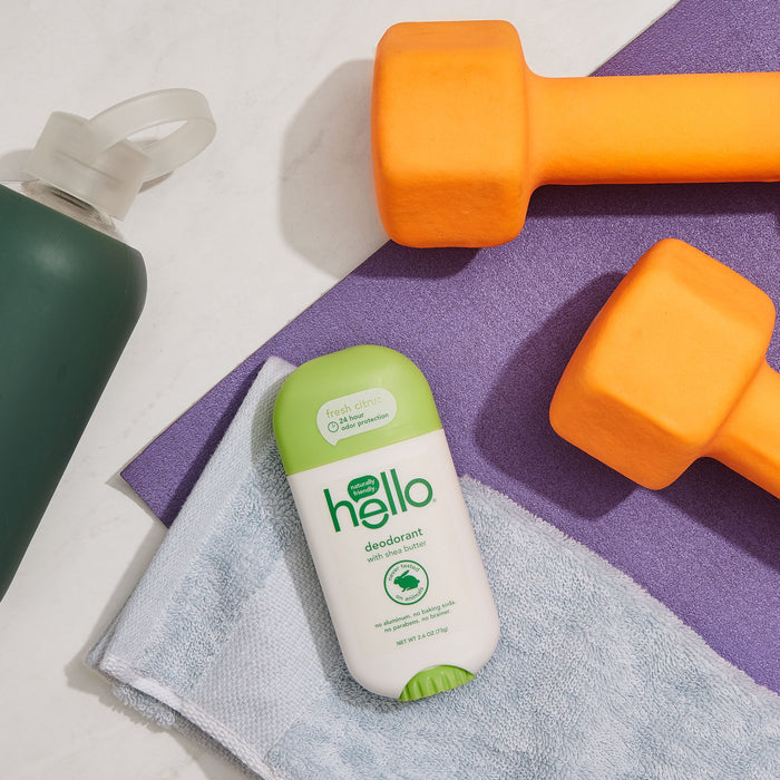 A flat lay features a green-capped hello Fresh Citrus Deodorant with Shea Butter on a light blue towel, alongside two orange dumbbells and a dark green water bottle, all set against a purple yoga mat.