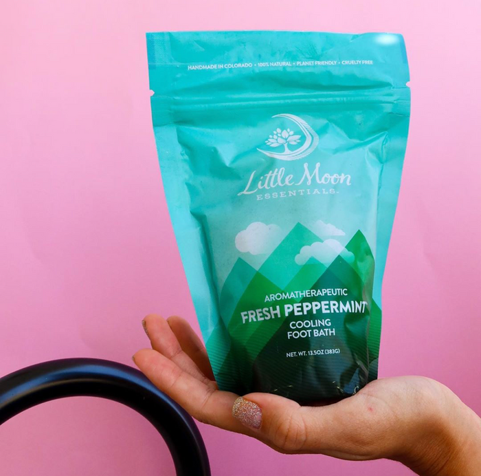 A hand holds a teal and green package of Little Moon Essentials Fresh Peppermint Foot Soak against a pink background, promising refreshing relief. The packaging highlights that it is handmade, 100% natural, and cruelty-free.