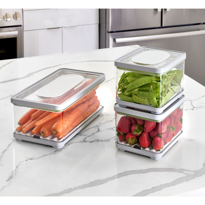 Three iDesign Fresh Produce Bins made from recycled plastic sit on the marble countertop, each holding carrots, peas, and strawberries. Featuring clear sides and white lids, they elegantly extend produce life against a backdrop of gray faucet and cabinets.